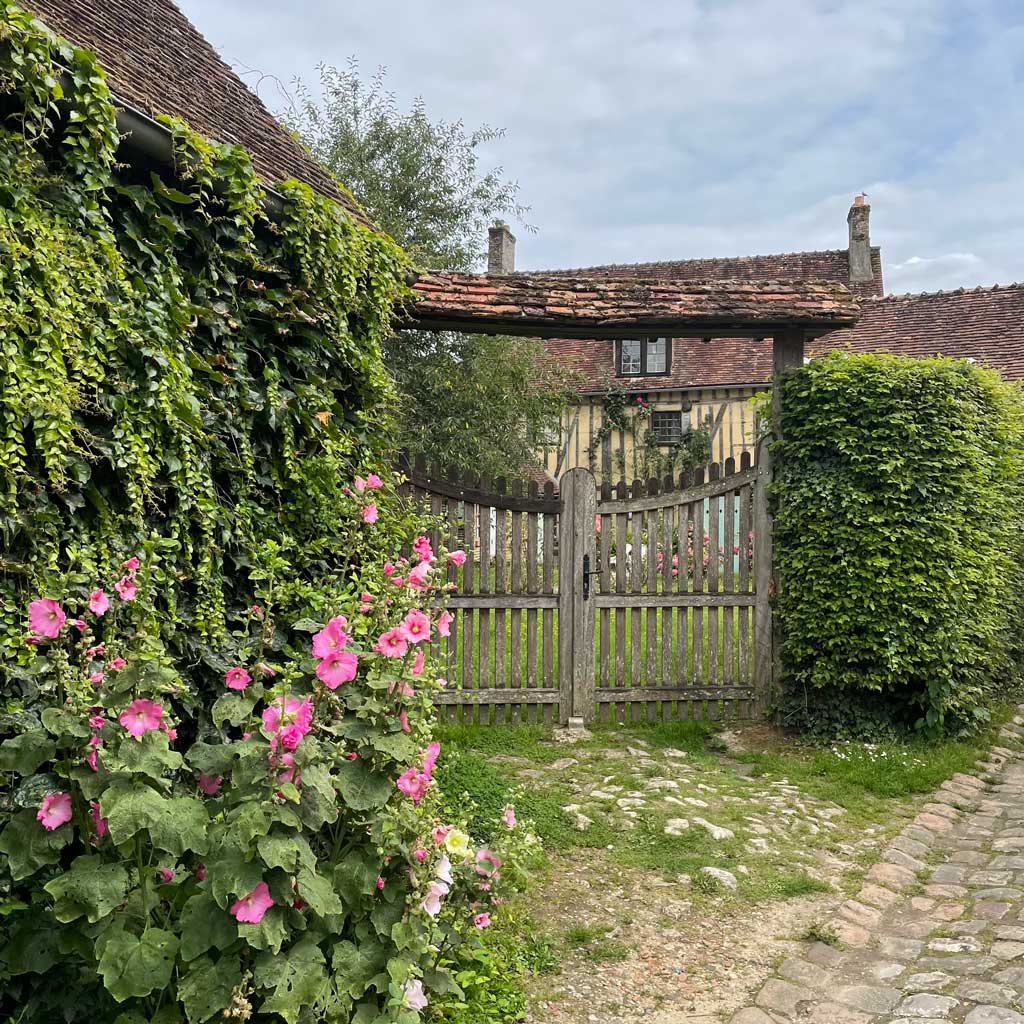 A slatted wooden gate under a tiled canopy and a stone path with pink hollyhocks and a house